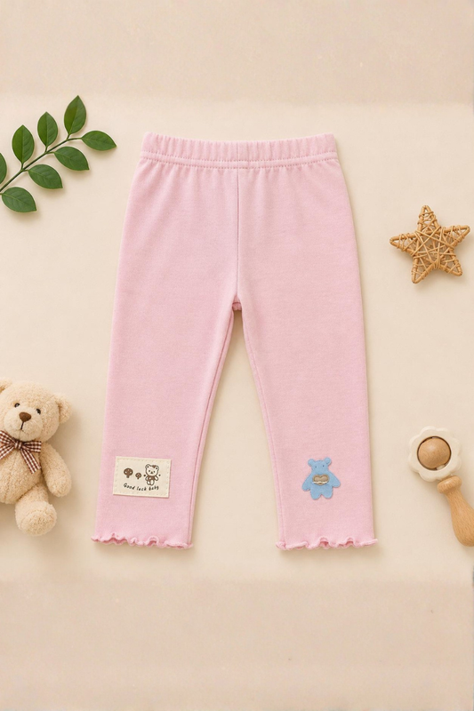 Pink baby leggings with a label and bear design on a beige background with toys and leaves.