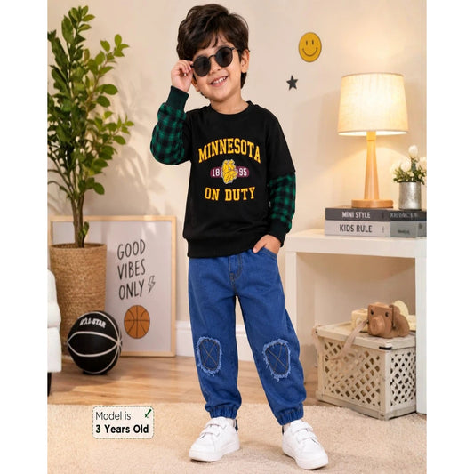 A 3-year-old boy modeling a black long-sleeve shirt with green plaid sleeves and yellow 'MINNESOTA 1895 ON DUTY' text, paired with blue jeans and white sneakers in a styled room setting.